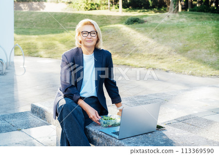 Business woman eating fast and healthy in front of laptop during job lunch break outdoors. Modern business lifestyle. Middle aged lady eats salad and use notebook pc on bench. Balanced diet lunch box. Business woman eating fast and healthy in front of laptop during job lunch break outdoors. Modern business lifestyle. Middle aged lady eats salad and use notebook pc on bench. Balanced diet lunch box. 113369590