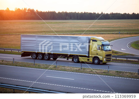 A truck with a tilt semi-trailer carries cargo along the highway in the evening against the backdrop of a summer sunset. The concept of cargo transportation, the logistics company, the work and rest A truck with a tilt semi-trailer carries cargo along the highway in the evening against the backdrop of a summer sunset. The concept of cargo transportation, the logistics company, the work and rest 113369959