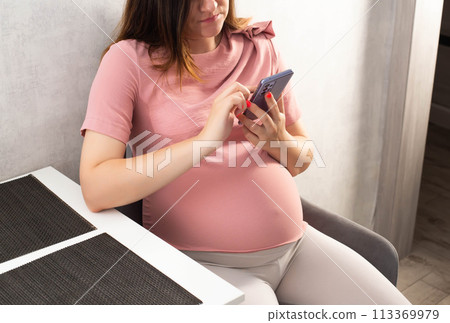 Pregnant girl with a mobile phone in the kitchen. The concept of the negative impact of phone radiation on the fetus in the womb. 113369979