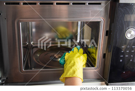 A girl's hand in a yellow glove with a sponge washes the microwave oven from grease and dirt. Foam and anti-grease. Cleaning household appliances in the kitchen 113370056