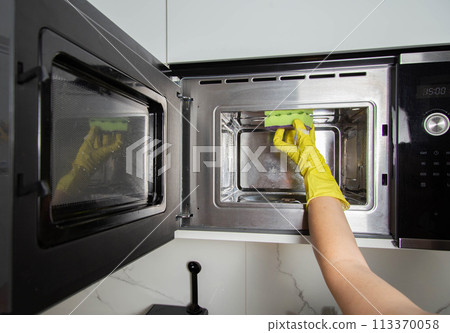 A girl's hand in a yellow glove with a sponge washes the microwave oven from grease and dirt. Foam and anti-grease. Cleaning household appliances in the kitchen A girl's hand in a yellow glove with a sponge washes the microwave oven from grease and dirt. Foam and anti-grease. Cleaning household appliances in the kitchen 113370058