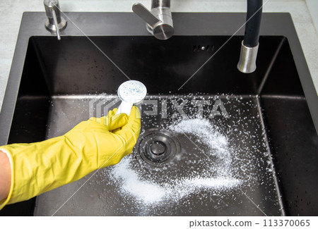 A man's hand in a yellow glove holds a spoon with oxygen bleach against the background of a kitchen sink. Cleaning from grease and dirt. A man's hand in a yellow glove holds a spoon with oxygen bleach against the background of a kitchen sink. Cleaning from grease and dirt. 113370065