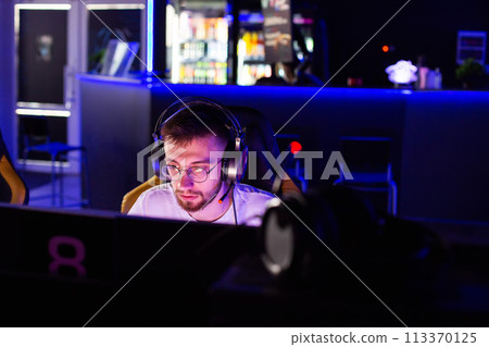 Adult bristly gamer in glasses headset plays video game on PC at computer club cyber sport tournament. Facial expressions. E-sport championship. Multiplayer online tactical shooter real-time strategy 113370125