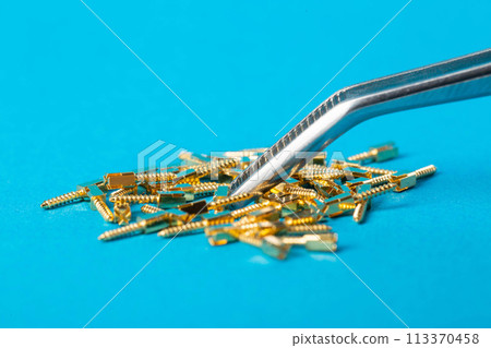 Metal dental pins on a blue background in tweezers in dentistry. Installation of pins for a dental crown in orthodontics, macro Metal dental pins on a blue background in tweezers in dentistry. Installation of pins for a dental crown in orthodontics, macro 113370458