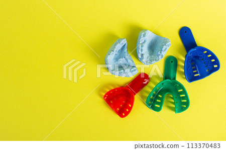 Blue plaster cast of a dental jaw and a dental tray for an impression of a dental jaw on a yellow background. The concept of orthodontics in dentistry, the manufacture of dentures and crowns. Copy Blue plaster cast of a dental jaw and a dental tray for an impression of a dental jaw on a yellow background. The concept of orthodontics in dentistry, the manufacture of dentures and crowns. Copy 113370483