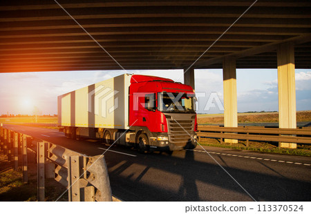 A red tractor with an isothermal refrigerated semi-trailer transports perishable goods with temperature conditions against the backdrop of sunset under the bridge. Delivery of perishable products to A red tractor with an isothermal refrigerated semi-trailer transports perishable goods with temperature conditions against the backdrop of sunset under the bridge. Delivery of perishable products to 113370524