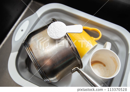 measuring spoon with granules of environmentally friendly oxygen bleach in a basin with dirty dishes. Cleaning grease and dirt from dishes, close-up 113370528