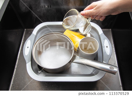 A woman's hand pours hot water into a basin with dirty dishes and environmentally friendly oxygen bleach. Washing dishes from grease and dirt. A woman's hand pours hot water into a basin with dirty dishes and environmentally friendly oxygen bleach. Washing dishes from grease and dirt. 113370529