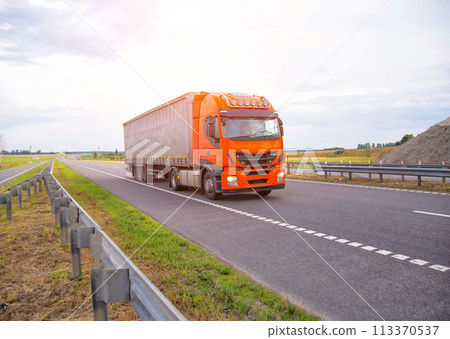 An orange truck with a semi-trailer curtain transports auto parts against the sky with clouds in the summer on the highway. Copy space for text, trucking 113370537