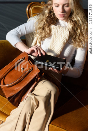 beautiful curly blond hair woman posing with a wallet and small shopper brown bag in a vintage chair 113371886
