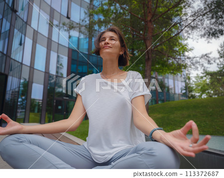 De-stress, unwind idea. peaceful female freelance employee practicing yoga exercises while sitting in bunch under glass skyscrapers after hard working day in office, holding hands in mudra De-stress, unwind idea. peaceful female freelance employee practicing yoga exercises while sitting in bunch under glass skyscrapers after hard working day in office, holding hands in mudra 113372687