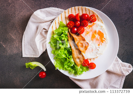 Close up of tacos with fried egg, bacon, tomato and lettuce on a plate on the table top view Close up of tacos with fried egg, bacon, tomato and lettuce on a plate on the table top view 113372739