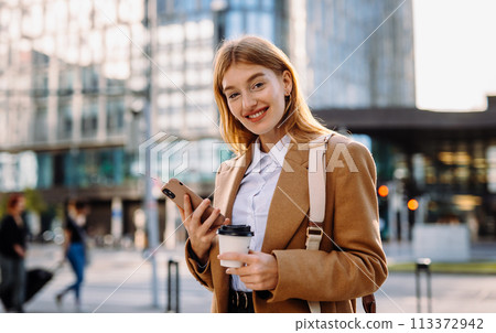 A woman in a blazer smiles at her phone in the city 113372942