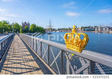 A golden crown sculpture adorns the Skeppsholmen Bridge under a blue sky, with Stockholms waterfront and ships in the background. Stockholm, Sweden 113373151