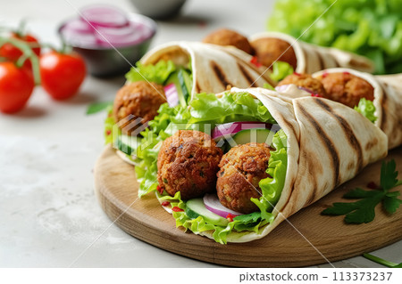 Delicious, fried falafel balls wrapped in pita bread with cucumbers, lettuce salad, red onion and sauce on wooden cutting board on white concrete background, perfect vegetarian snack 113373237