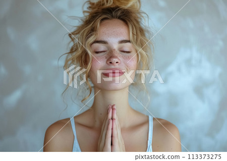 Blonde girl with curly hair doing in meditation the day with eyes closed, hands in prayer position, her face is calm and peaceful 113373275