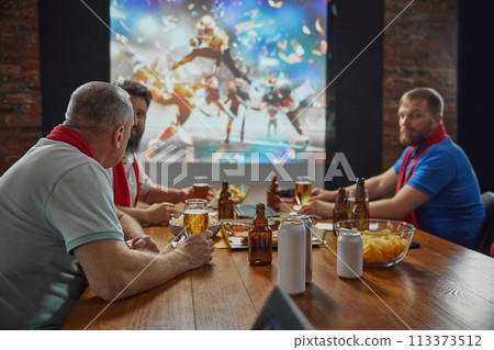 Friends coming together to watch an online American football match on TV, with beer and snacks, expressing enjoyment of the game. 113373512