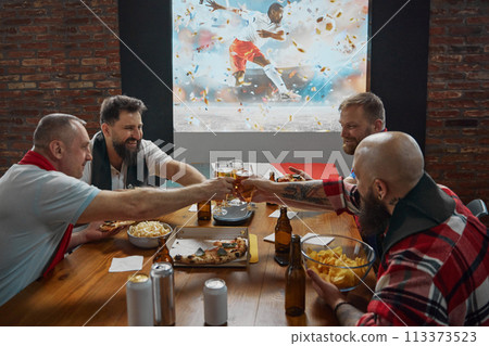 Cheers. Friends, men sitting ta table and watching online football match translation, clinking beer glasses for scoring goal. 113373523