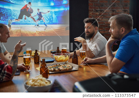 Friends sitting together, immersed in the excitement of an online football match broadcast on TV, with beer and snacks on table. Game discussion 113373547