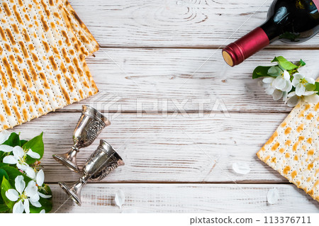 Jewish religious holiday of Passover. Bottle of wine, matzo and flowers on white wooden background. Jewish religious holiday of Passover. Bottle of wine, matzo and flowers on white wooden background. 113376711
