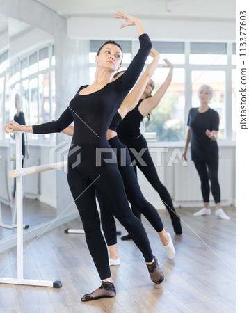 Mature teacher conducts class with girls in ballet studio, observes doing Batman exercise. 113377603