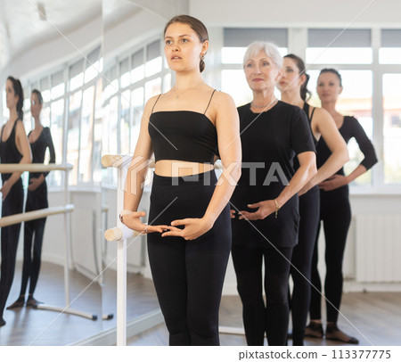 Women stand in fifth position near ballet barre during group training in dance studio Women stand in fifth position near ballet barre during group training in dance studio 113377775