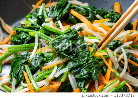 A cooking scene in which vegetables (spinach, carrots, and bean sprouts) are stir-fried in a frying pan. 113378712