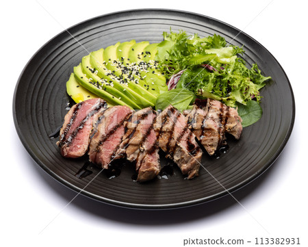 grilled cooked piece of tuna fillet with sliced avocado and salad on black ceramic plate 113382931