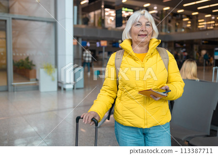 Airport terminal. Happy traveling Caucasian 60s woman is waiting at the departure gate to board the plane, using mobile smartphone and checking destination online.  113387711