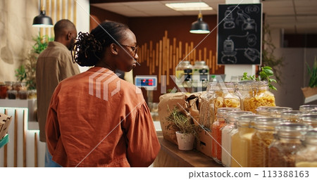 African american client examines bio homemade bulk products, looking to buy natural ethically sourced food alternatives for healthy nutrition. Woman shopping for goods at zero waste store. Camera 2. African american client examines bio homemade bulk products, looking to buy natural ethically sourced food alternatives for healthy nutrition. Woman shopping for goods at zero waste store. Camera 2. 113388163