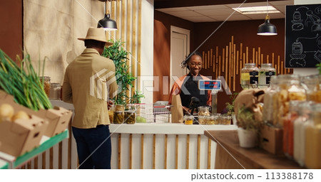 African american seller working at supermarket cash register counter, weighting organic additives free fruits and vegetables. Vendor selling fresh bio produce to male customer at checkout. Camera 2. 113388178