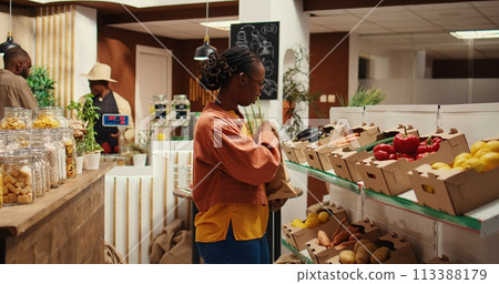 Vegan person searching for freshly harvested vegetables from crates, shopping for natural groceries at local zero waste eco store. African american client looking at organic food items. Camera 2. Vegan person searching for freshly harvested vegetables from crates, shopping for natural groceries at local zero waste eco store. African american client looking at organic food items. Camera 2. 113388179