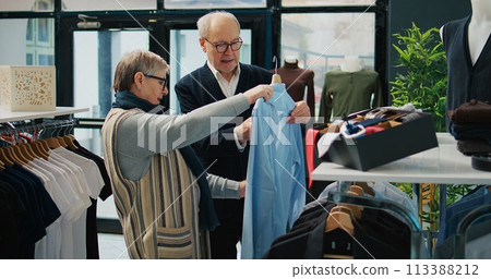 Senior couple shopping for formal outerwear at fashion boutique, looking for trendy elegant clothing items to create new outfits for elderly man. People searching for stylish merchandise. Camera B. Senior couple shopping for formal outerwear at fashion boutique, looking for trendy elegant clothing items to create new outfits for elderly man. People searching for stylish merchandise. Camera B. 113388212
