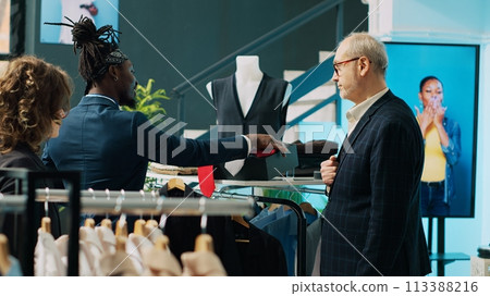 Fashion stylist making trendy recommendations for senior client, pairing an expensive tie to match a black elegant shirt. African american worker selling designer items to man. Camera A. 113388216