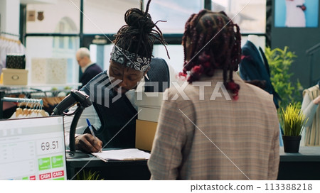 African american deliveryman receiving requested clothes packed in boxes, arriving at fashion showroom to pick up order and sign papers. Woman employee preparing items at cash register. Camera A. 113388218