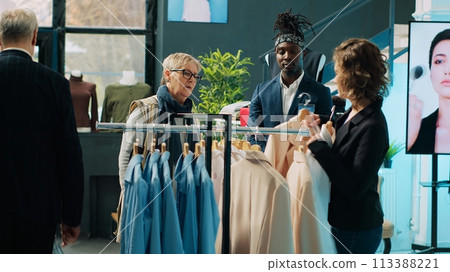 Fashion stylist suggesting clothing items to regular customer, team of store workers help elderly woman to buy trendy clothes on sale. Retail clerks assisting client during shopping spree. Camera A. 113388221