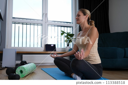 Young woman sitting in lotus position on rubber mat at home Young woman sitting in lotus position on rubber mat at home 113388518