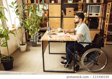 Art therapy. Young positive man sitting on wheelchair at home in living room and drawing. Relaxation 113390195