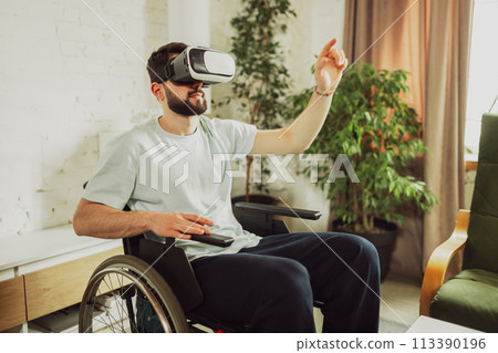 Young man sitting on wheelchair in living room at home, wearing VR glasses and exploring news innovative technology 113390196