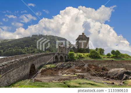 Waterside Eilean Donan Castle with stone bridge, fortified walls under dramatic sky 113390610