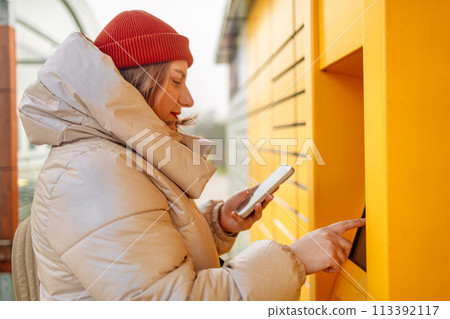 Smiling beautiful Caucasian woman client holding cardboard boxes and hands show smartphone using automated self service post terminal machine or locker. Mail shipping concept.  113392117
