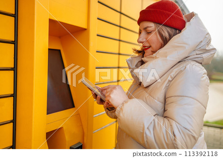 Happy smiling woman client with box at outdoor automated parcel machine choosing operation on touch screen by smart phone  113392118