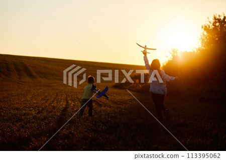 Running boy and girl holding two yellow and blue airplanes toy in the field during summer sunset. Kids dreams of flying and aviation. Running boy and girl holding two yellow and blue airplanes toy in the field during summer sunset. Kids dreams of flying and aviation. 113395062