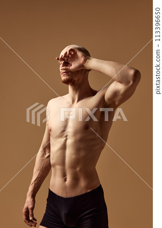 Shirtless muscular man shielding eyes from light against beige studio background. Young model with toned body. Shirtless muscular man shielding eyes from light against beige studio background. Young model with toned body. 113396650
