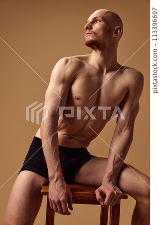 Young athletic man posing in underwear on stool looking away against sandy studio background. Young athletic man posing in underwear on stool looking away against sandy studio background. 113396697