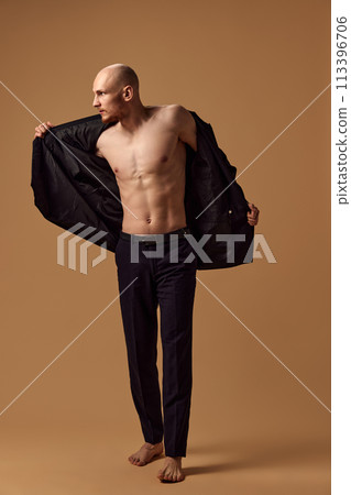 Shirtless young toned man dressed elegant trousers and jacket showing casual sophistication against sandy studio background. Shirtless young toned man dressed elegant trousers and jacket showing casual sophistication against sandy studio background. 113396706