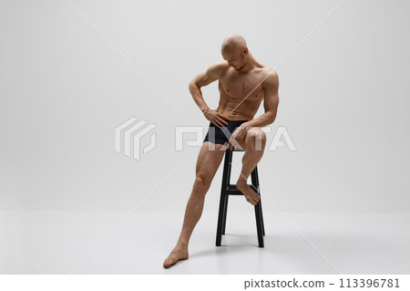 Athletic male model posing naked wearing in black underwear sitting on stool against grey studio background. 113396781