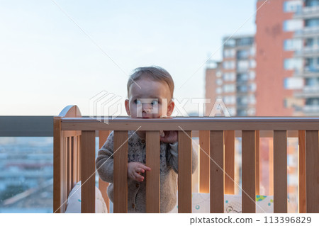 Baby in a crib against the backdrop of tall city buildings. Urban environment concept 113396829