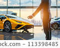 Elegant man in a suit pointing at a yellow luxury sports car in a showroom 113398485