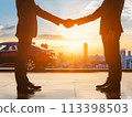 Silhouette of two businessmen shaking hands with a team in the background, set against a vibrant sunset 113398503